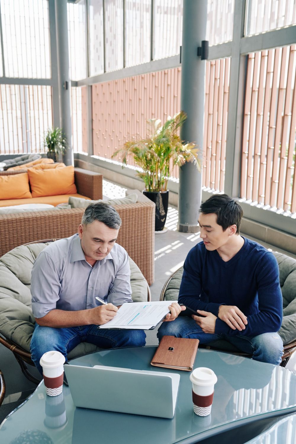 Two men review documents seated near a laptop. One writes, the other holds a tablet, in a modern lounge.