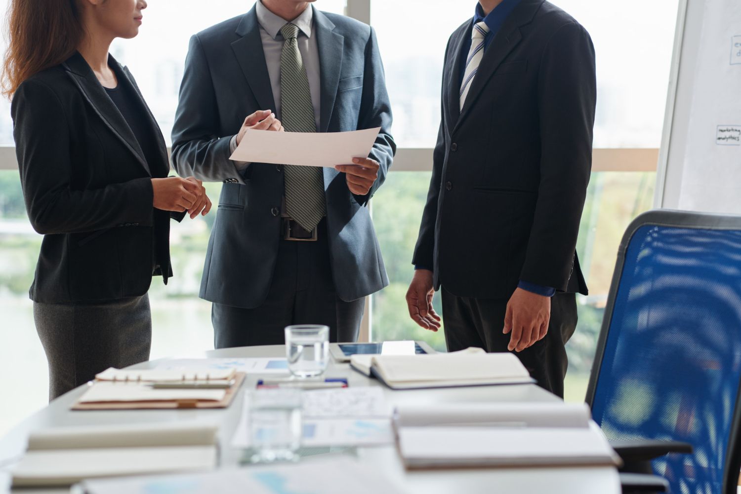 Three people in business suits reviewing documents at a conference table, sunlight.