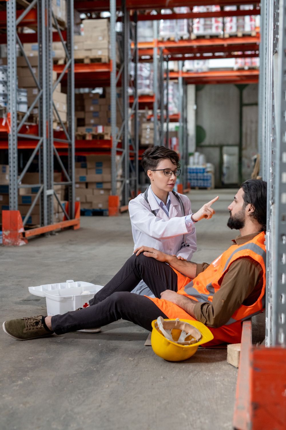 Warehouse worker seated, examined by a person in a lab coat, pointing, yellow hard hat nearby.