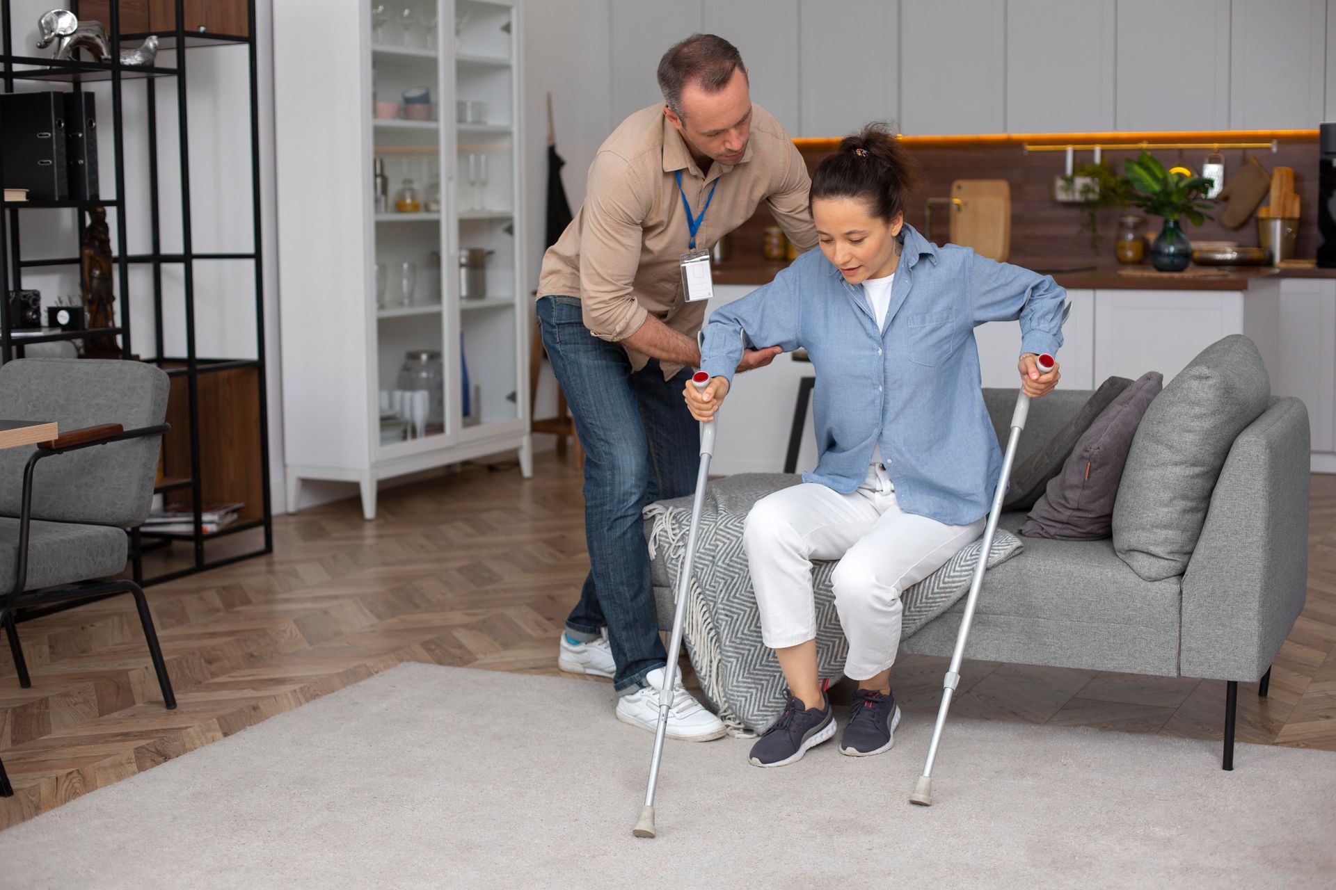 Man helps person with crutches sit on a couch in a living room.