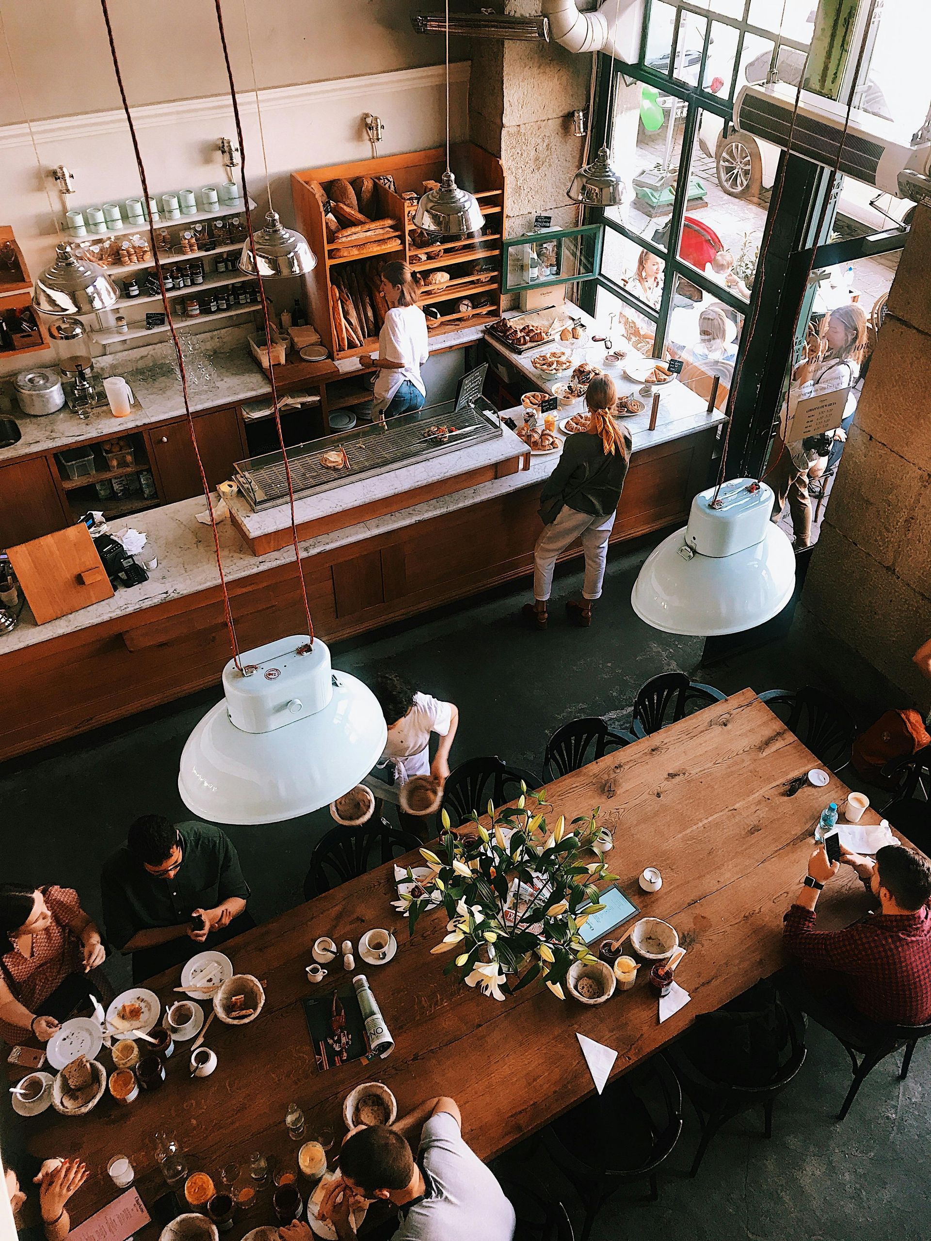 High-angle view of a cafe interior. Customers dine at a large wooden table under hanging lights, near a counter with baked goods.
