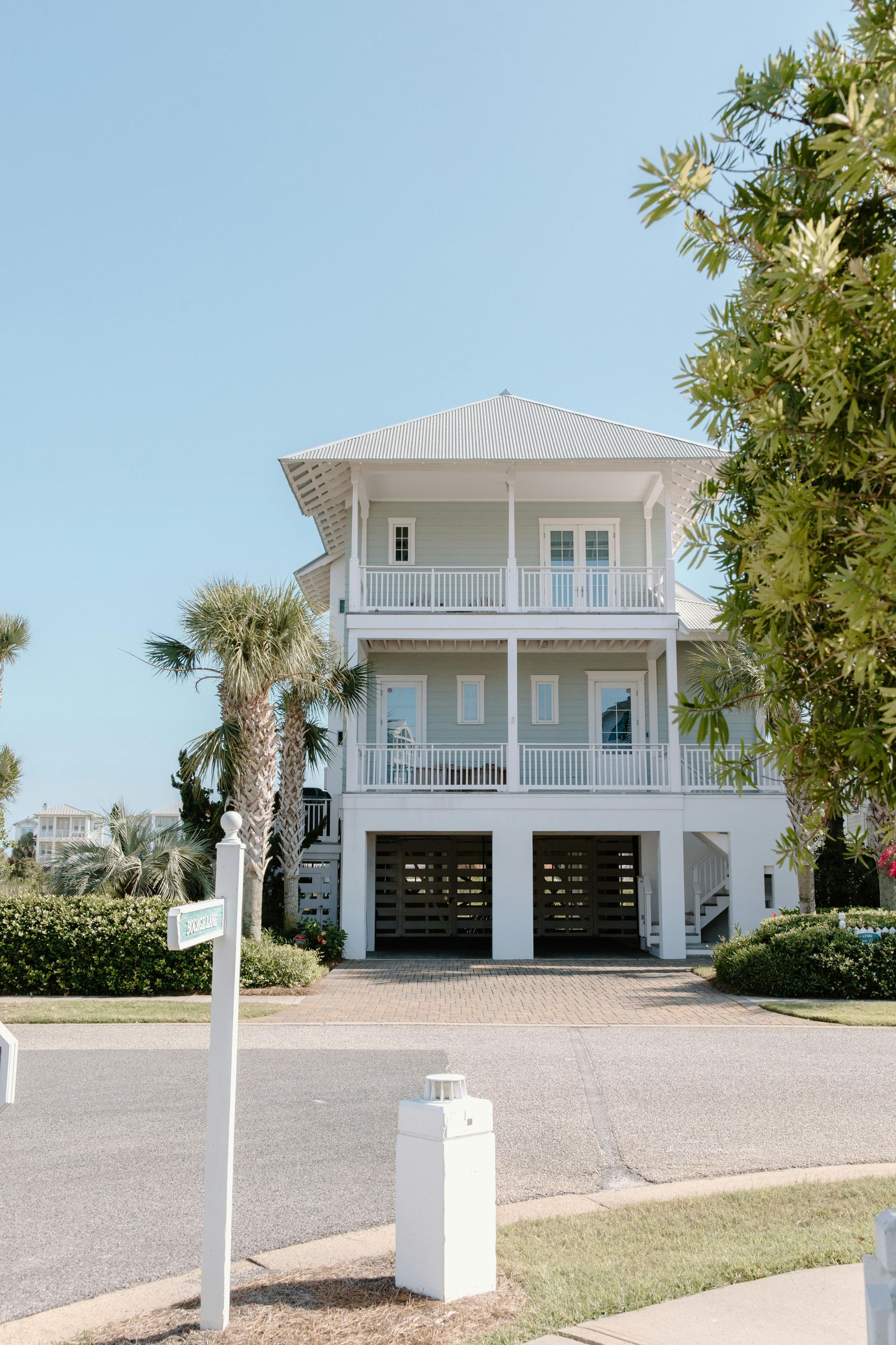Two-story beach house, light blue exterior, white railings, carport below, clear blue sky.