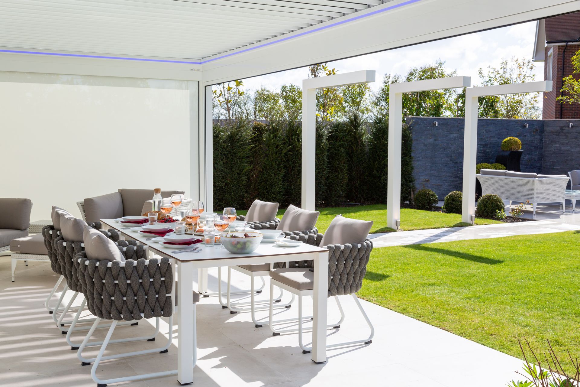 Outdoor dining table set under a white pergola with seating for eight, overlooking a lawn.
