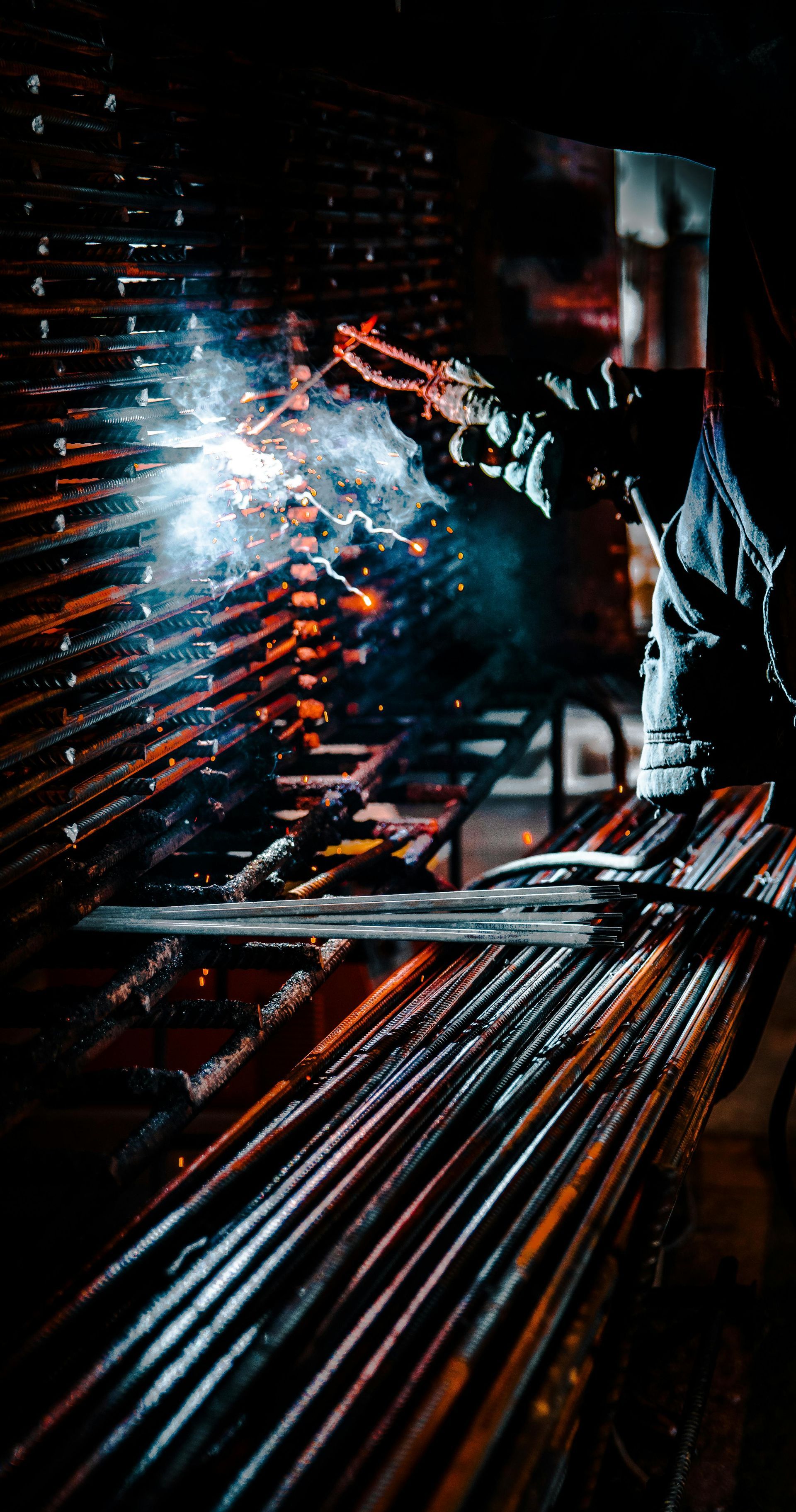 Worker welding metal in a dark workshop, bright sparks flying over red-lit machinery.