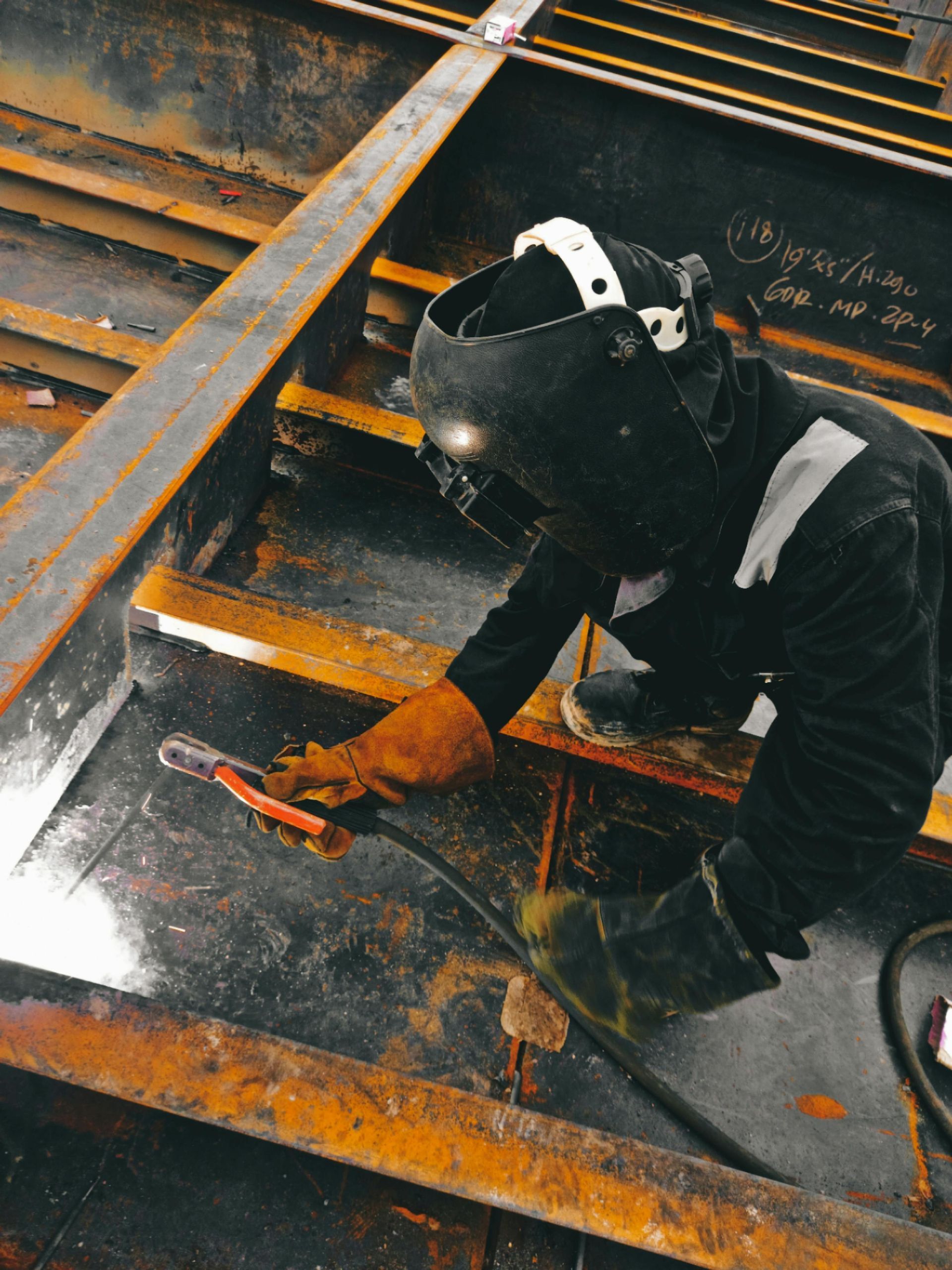 Worker welding metal beams on a steel structure, wearing protective gear and gloves, with orange sparks visible