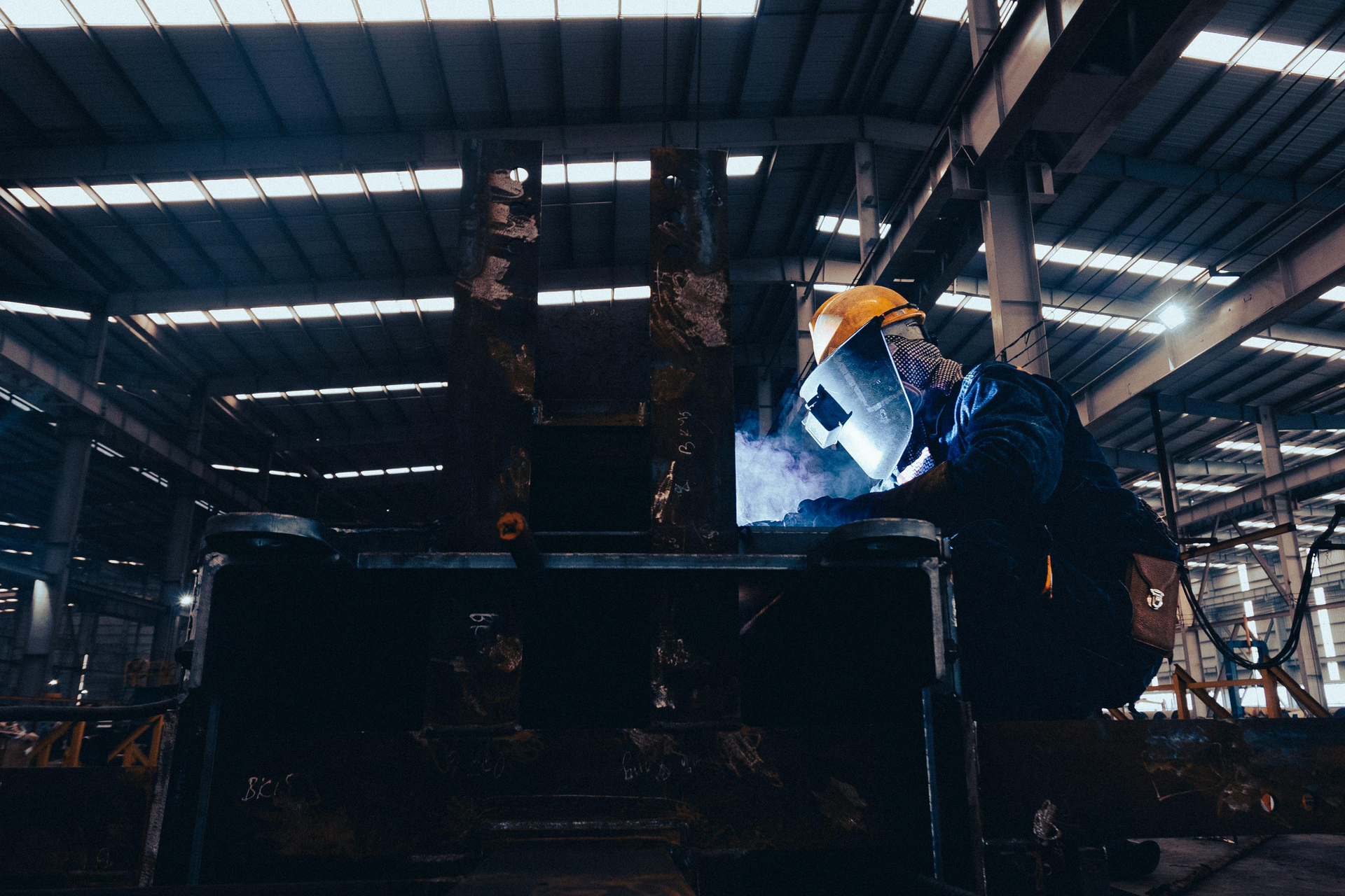 Worker welding a large metal structure inside a dim industrial factory, bright sparks glowing.