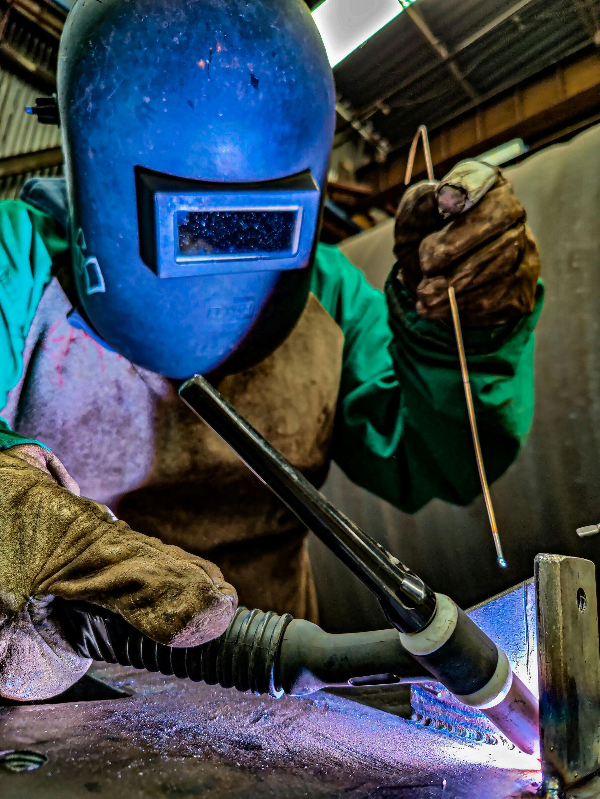 Welder in blue helmet and green gloves using a torch on metal, sparks flying in a workshop