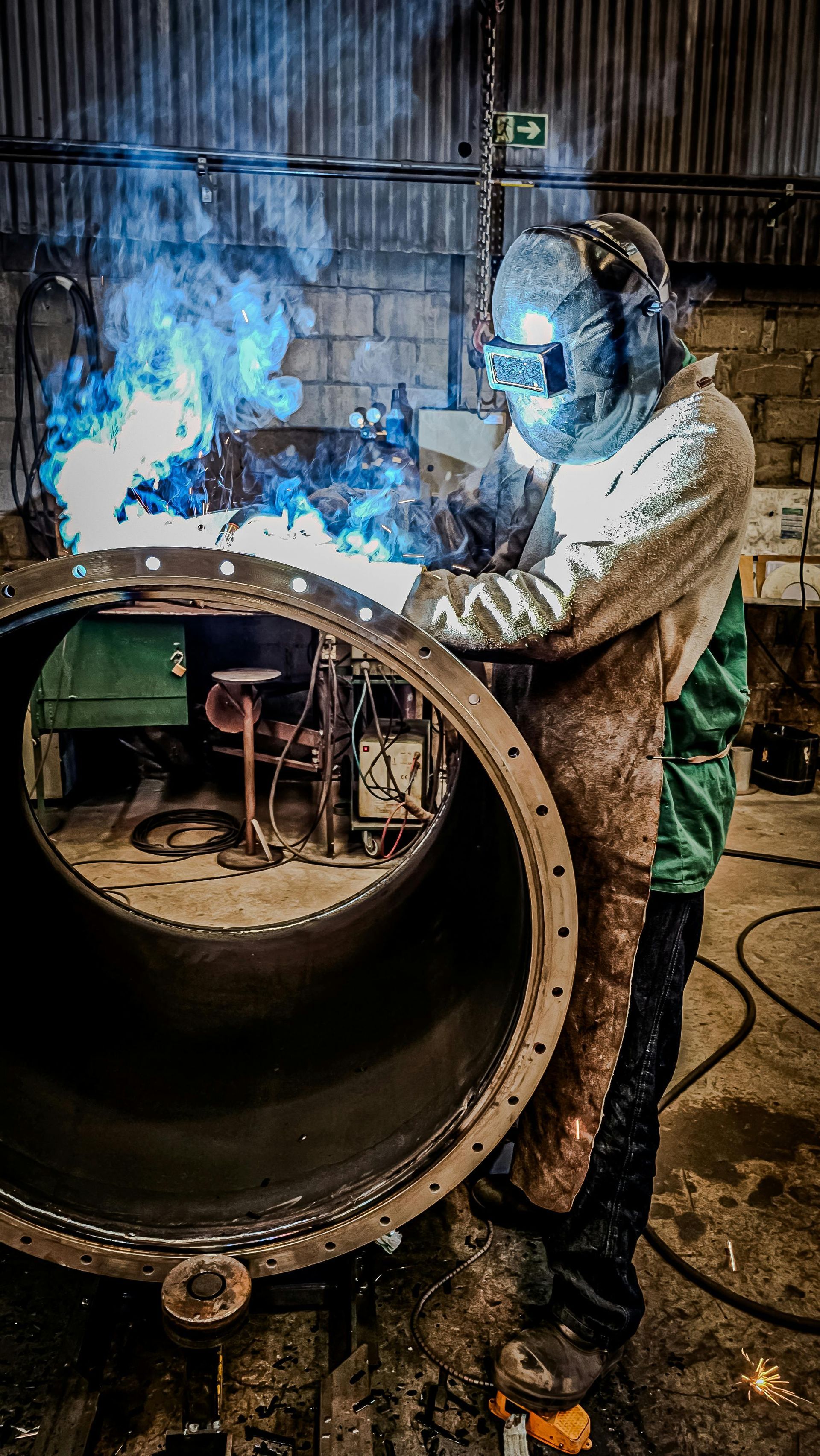 Welder in protective gear working on a large metal pipe, with bright blue sparks in an industrial setting