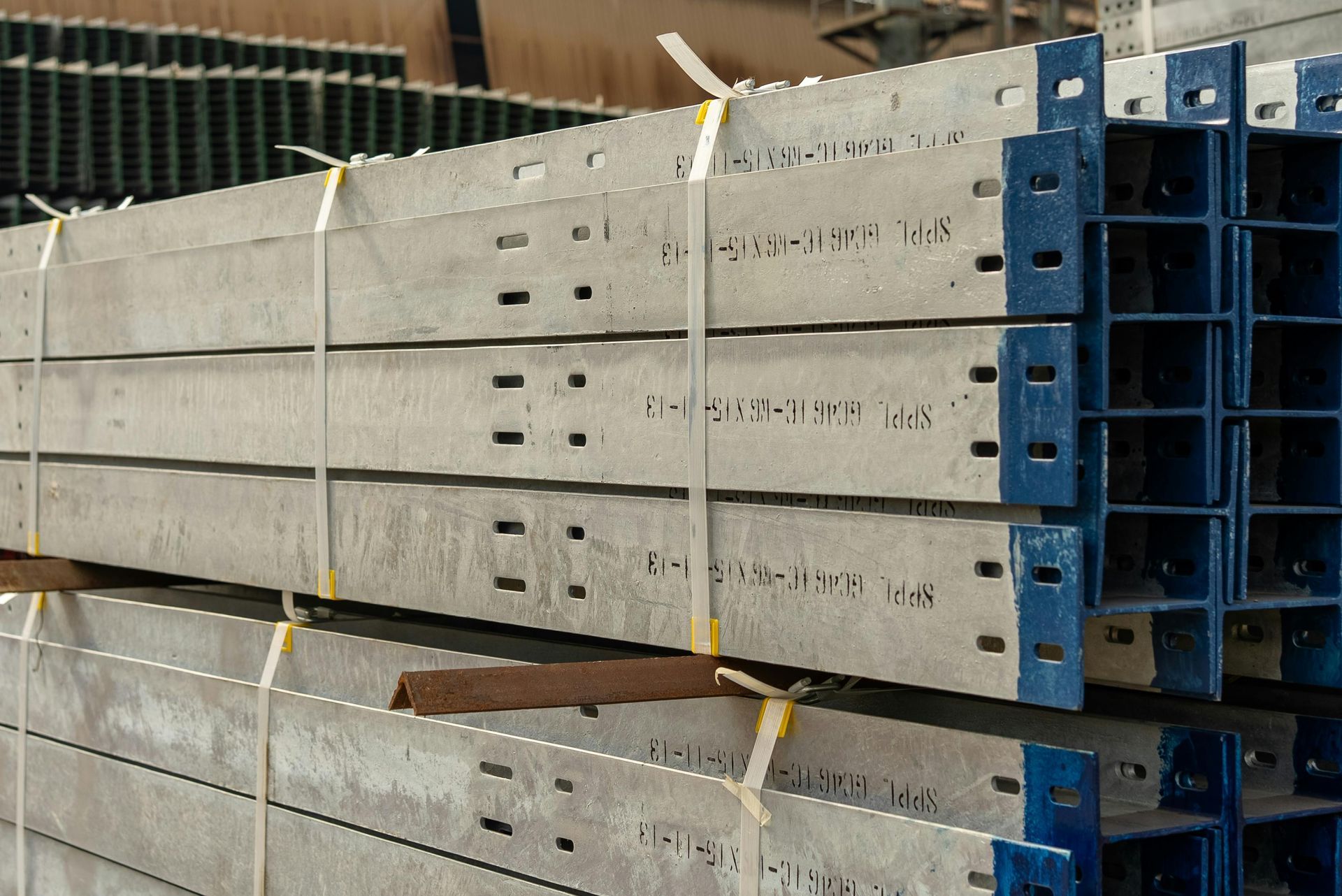 Stacked blue-gray metal scaffolding beams secured with yellow straps in a warehouse.