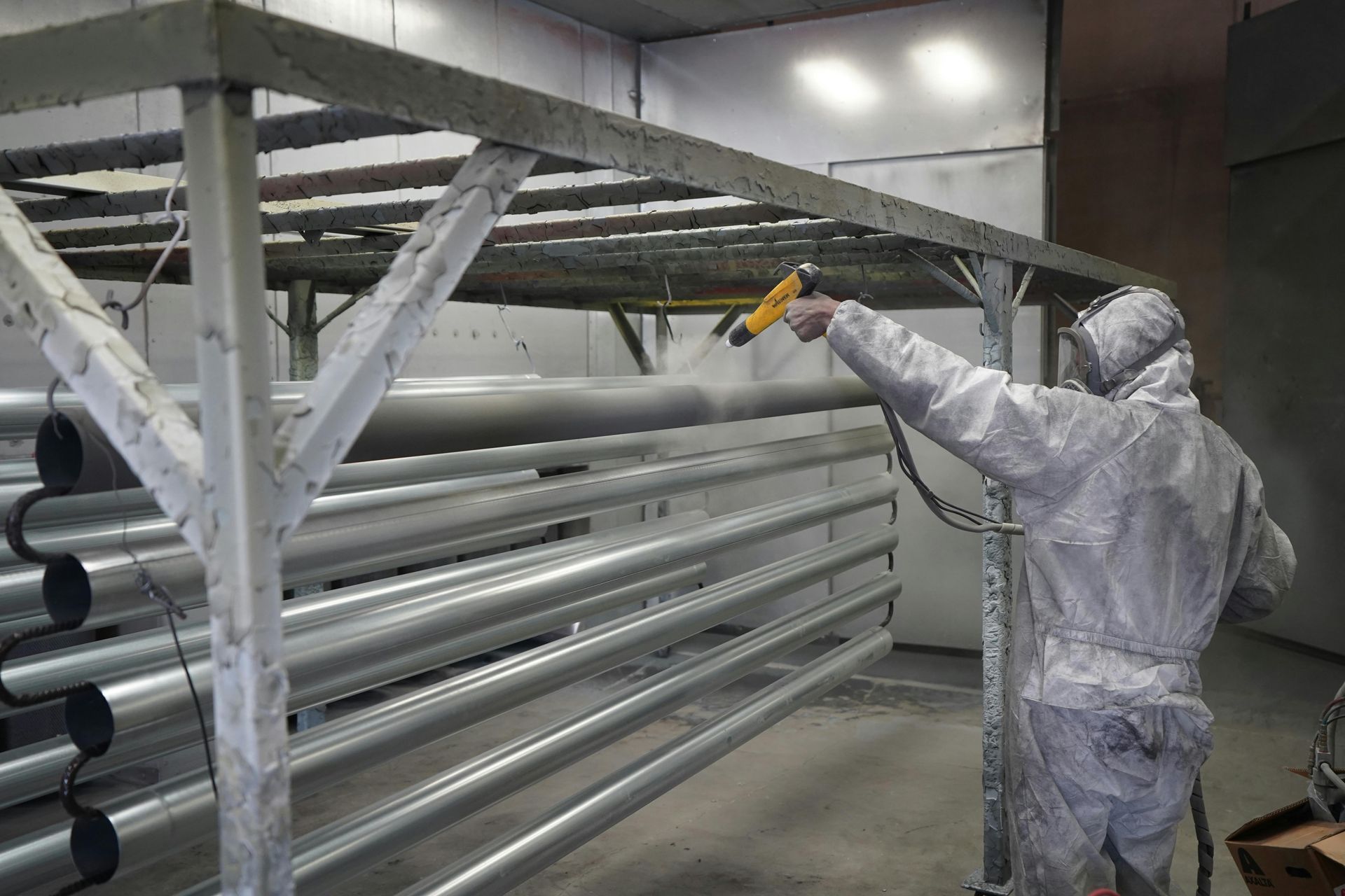 Worker in white protective suit spraying paint on large metal pipes in an industrial workshop