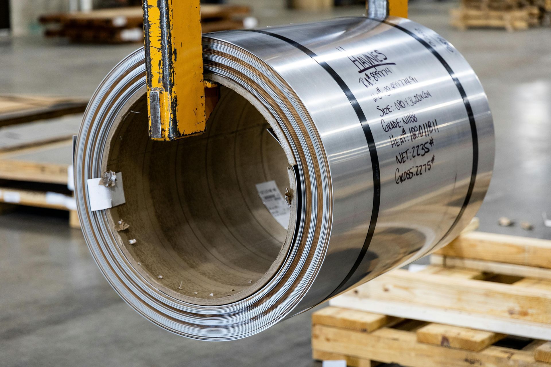 Coiled silver metal tubing with a yellow forklift hook lifting it in a warehouse or factory yard
