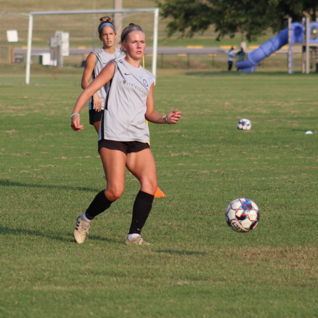 Woman in soccer uniform dribbling ball on a grassy field. Another player in background.