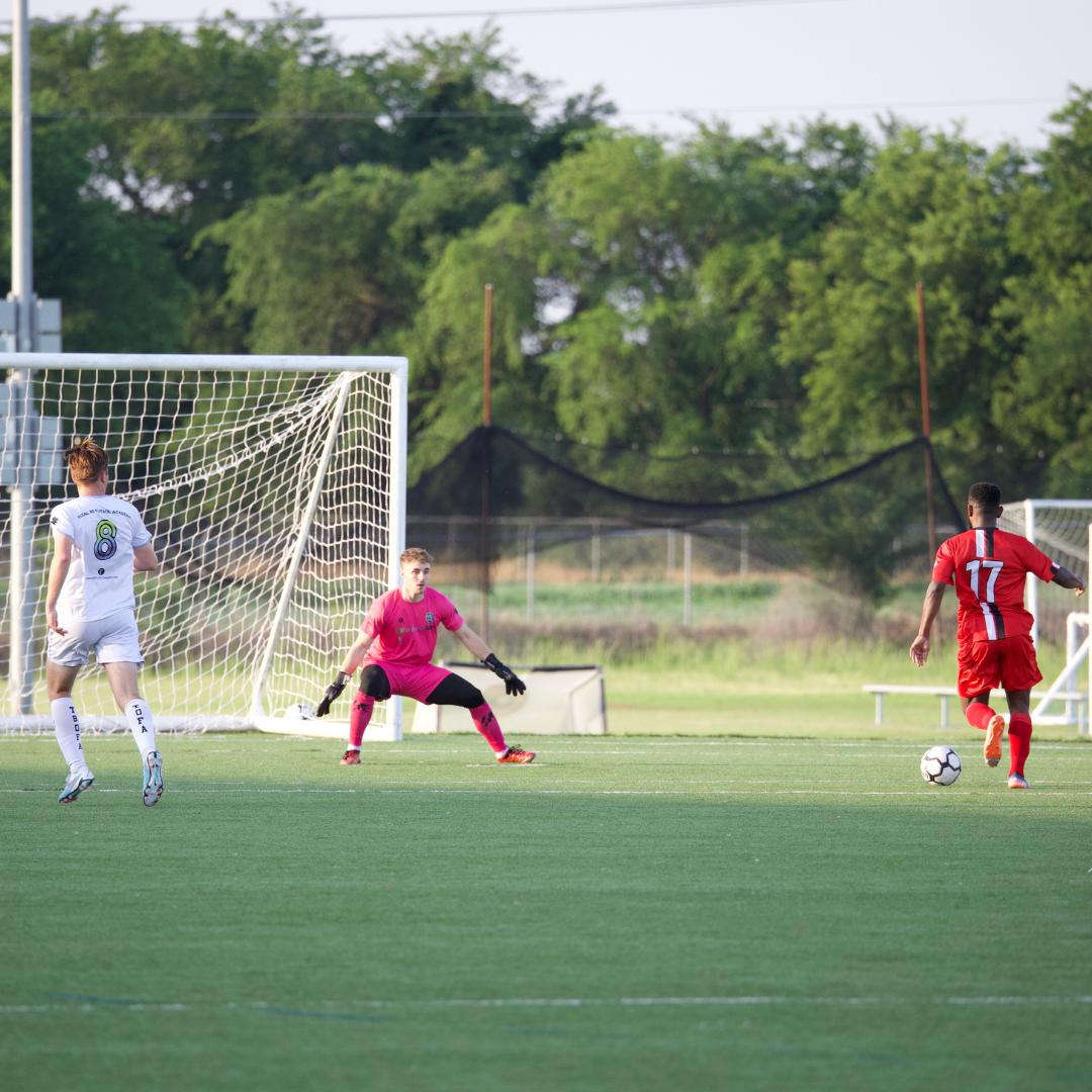 Soccer game: Goalie in pink suit defends net as red-shirted player dribbles ball. A white-shirted player runs nearby.