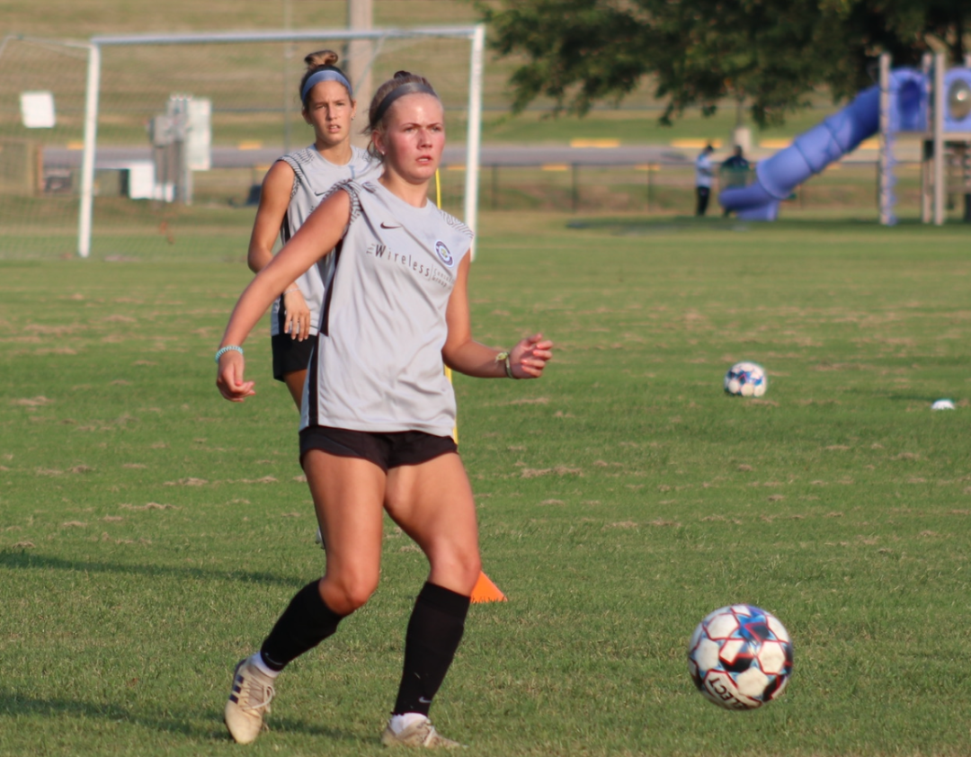 Soccer player in grey top dribbles ball on green field, another player in background.