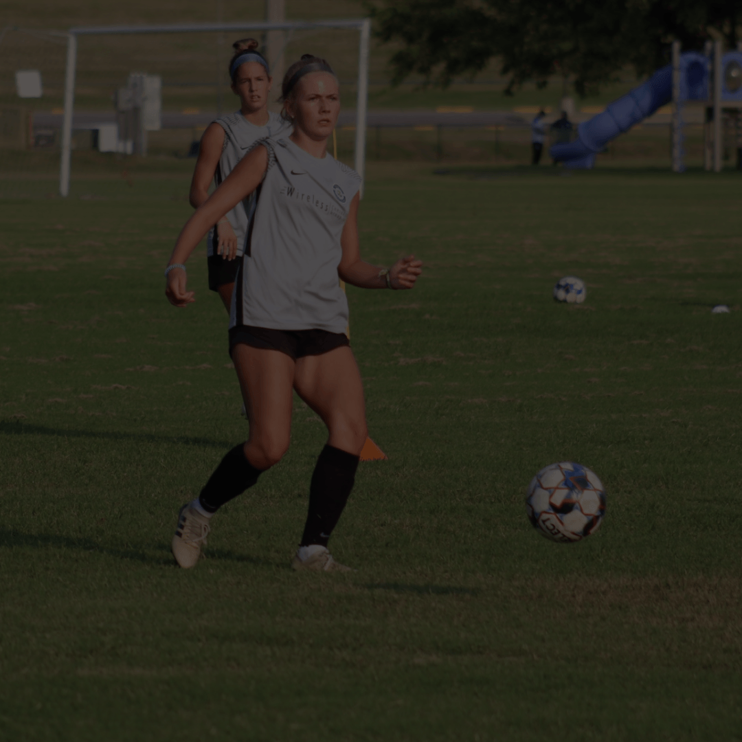 Woman in a grey shirt kicks a soccer ball on a field. Another woman is in the background.