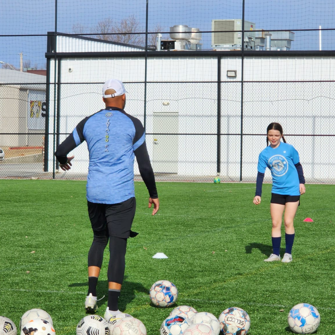 A soccer coach instructs a young girl on a field. Balls are in front.