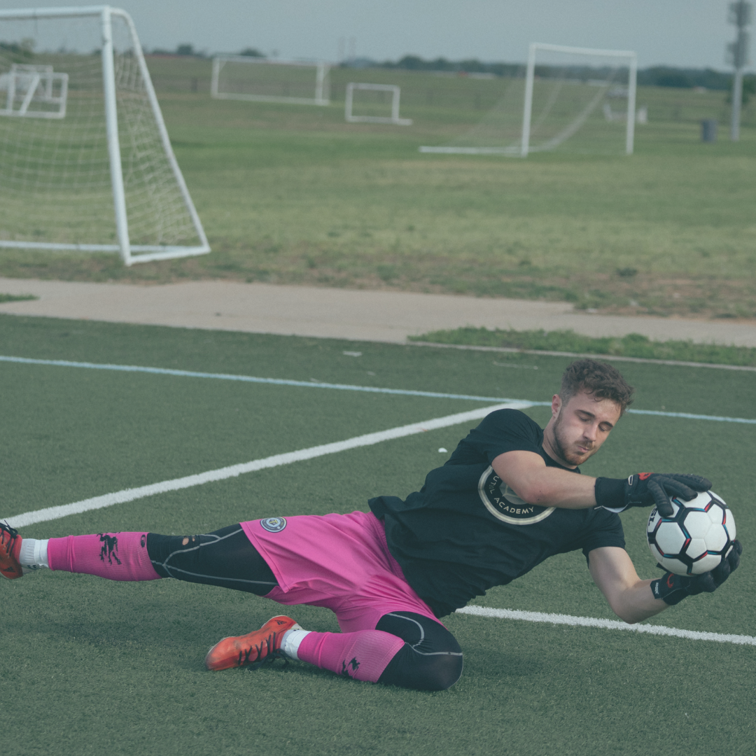 Soccer goalie in pink, diving to catch the ball on a green field.