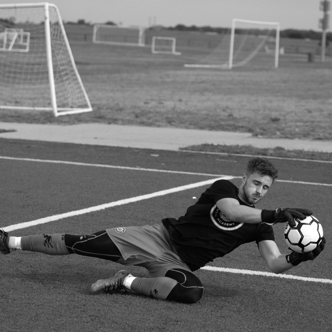 Soccer goalie dives to catch the ball during a game on a field.