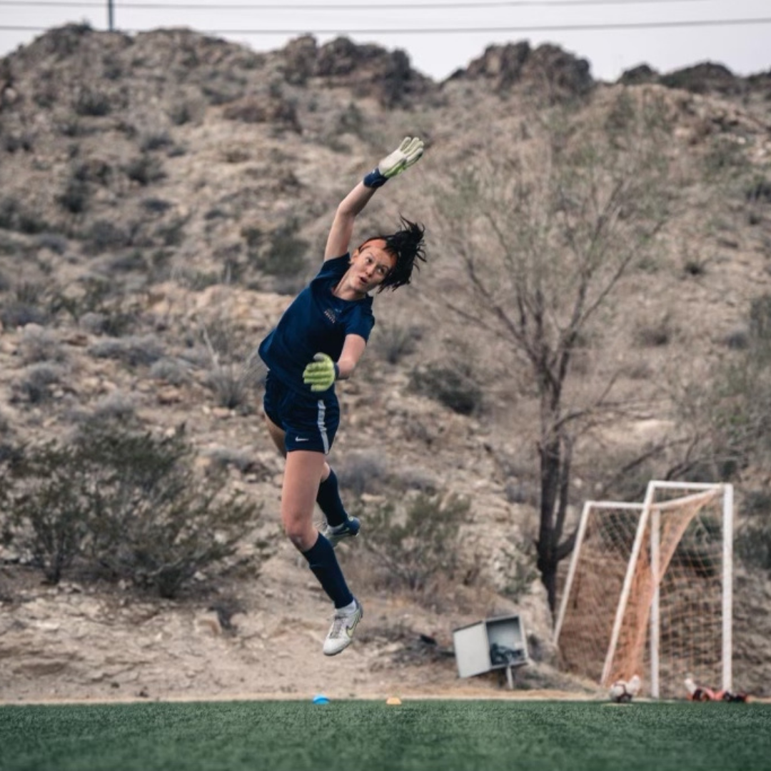 Soccer goalie in mid-air, diving for the ball, wearing navy blue uniform, set in a desert environment.
