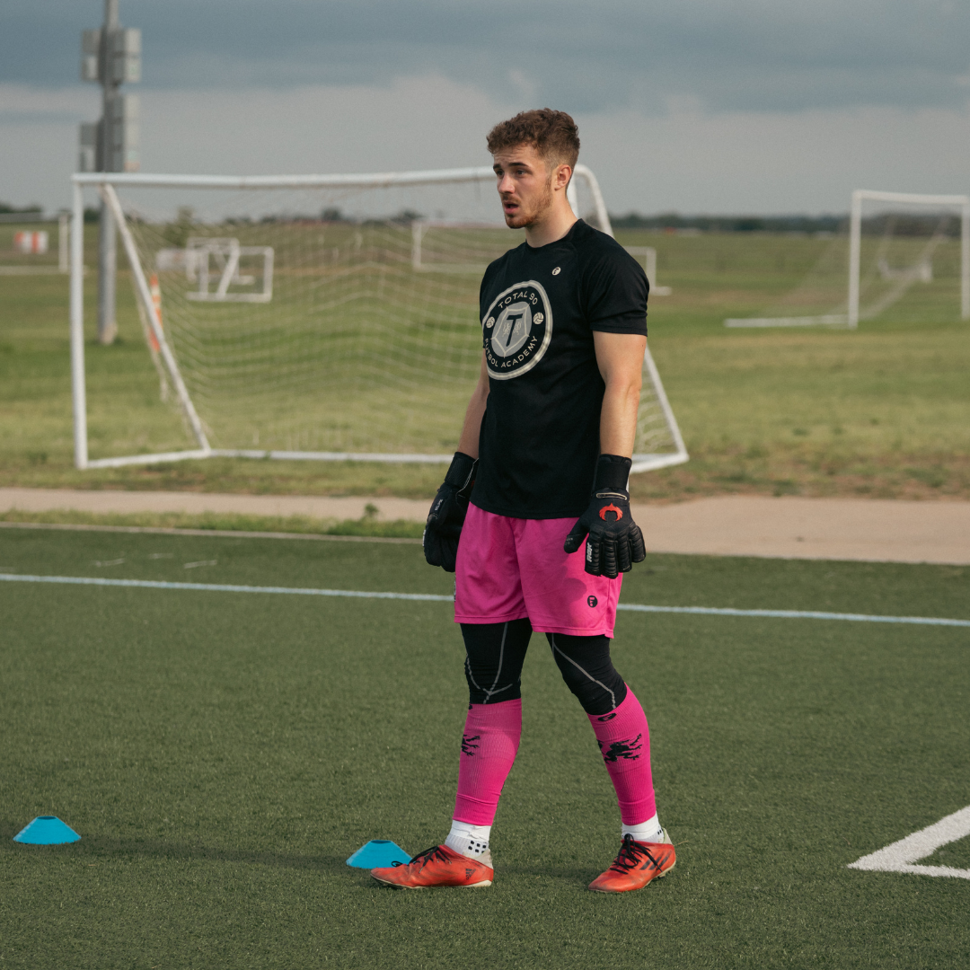 Soccer goalie in pink shorts and socks, black gloves, and tee shirt on a field.