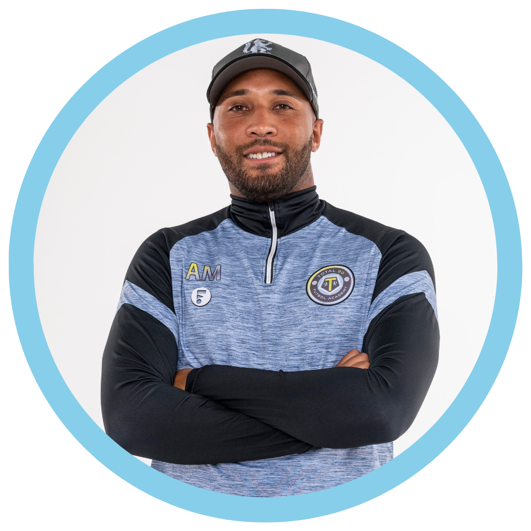 Man with arms crossed, wearing a cap and team shirt, smiling in front of a light background.