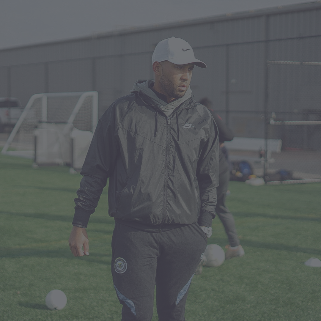Man in athletic wear on a soccer field, wearing a cap, looking on.