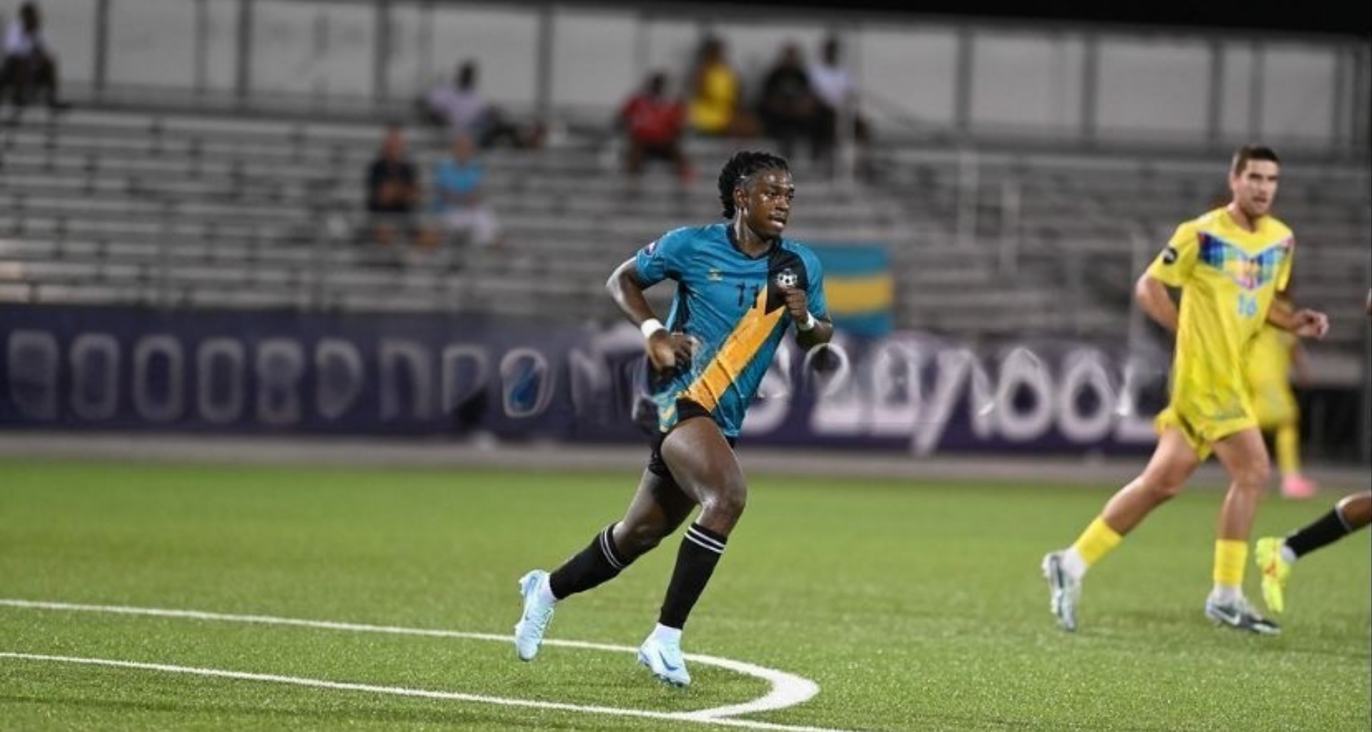 A soccer player in a teal and yellow jersey runs on a green field during a night game.