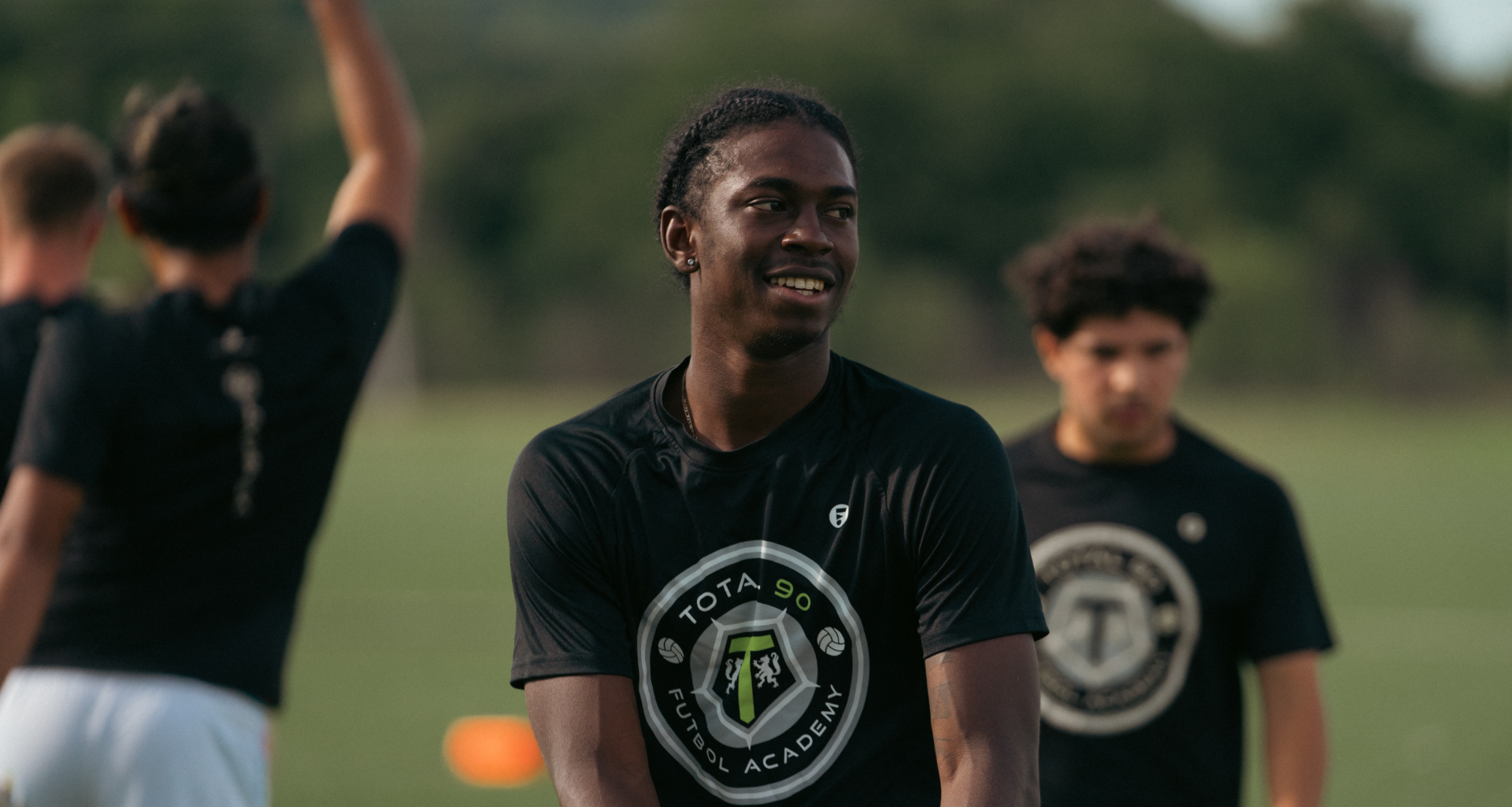 Smiling Black man in black shirt at soccer field, surrounded by teammates.