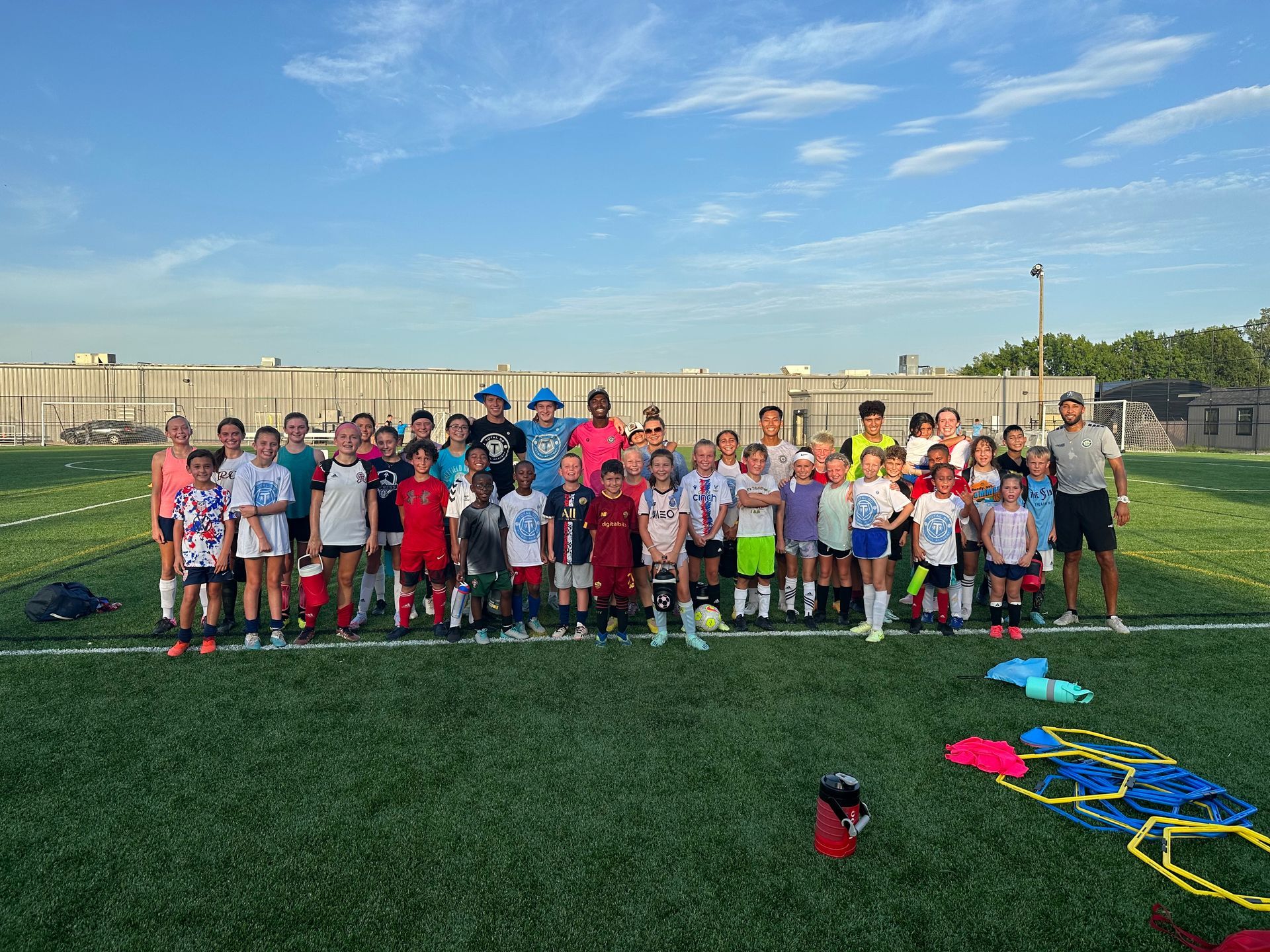 Group of kids in soccer gear on a green field, posing for a photo under a blue sky.