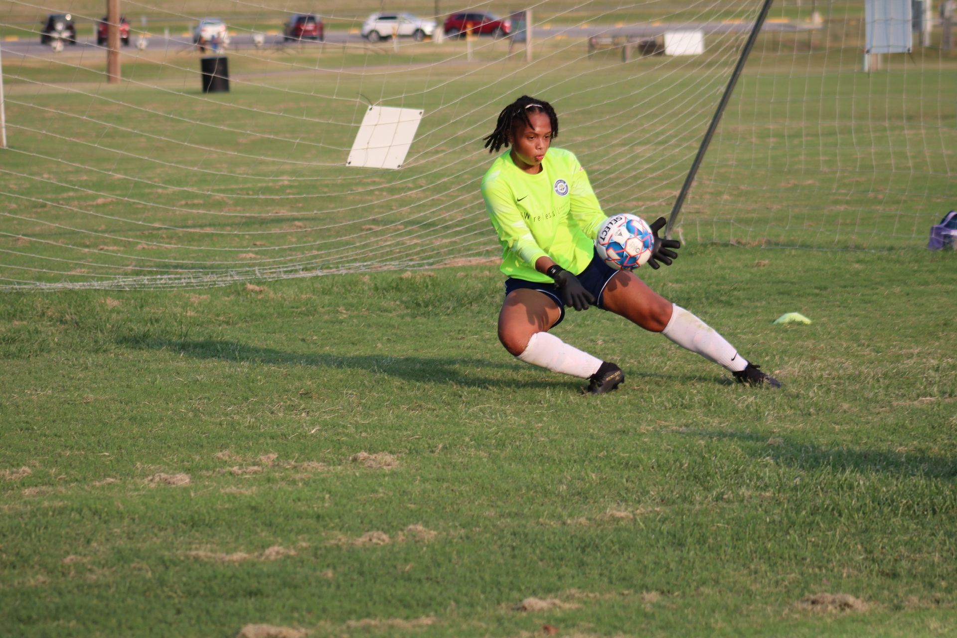 Soccer goalie dives to catch the ball in a green jersey and black shorts on a grassy field.