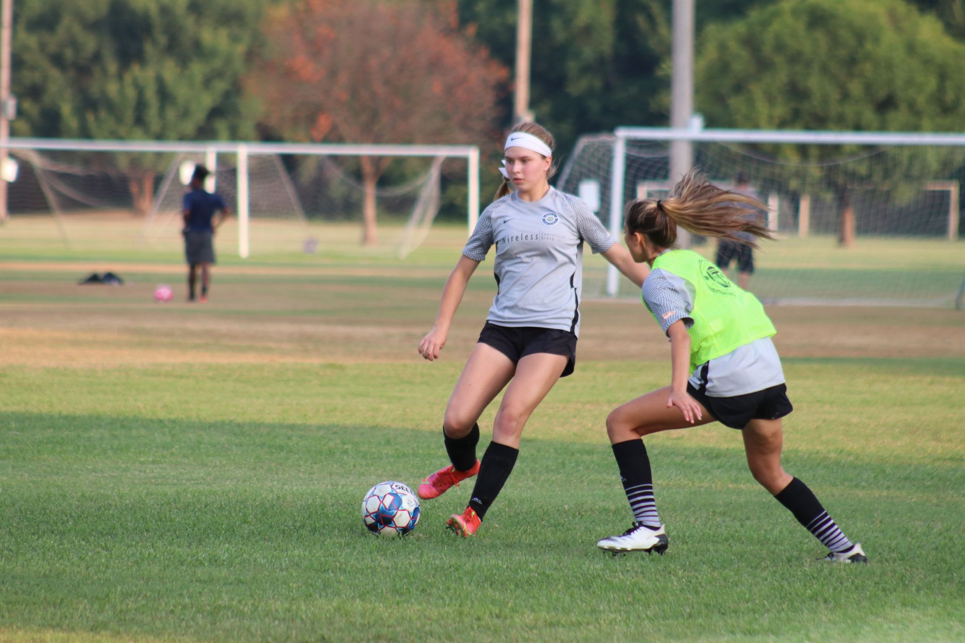 Soccer players on a field. One dribbles the ball, while another defends. Goals and trees in the background.