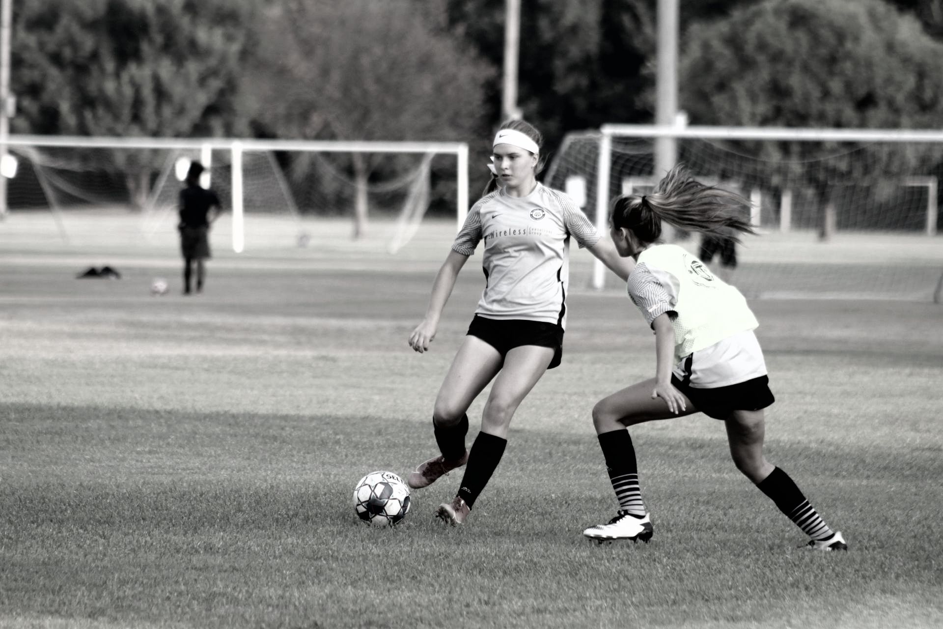 Two young women playing soccer on a field; one dribbles the ball, the other attempts to defend.