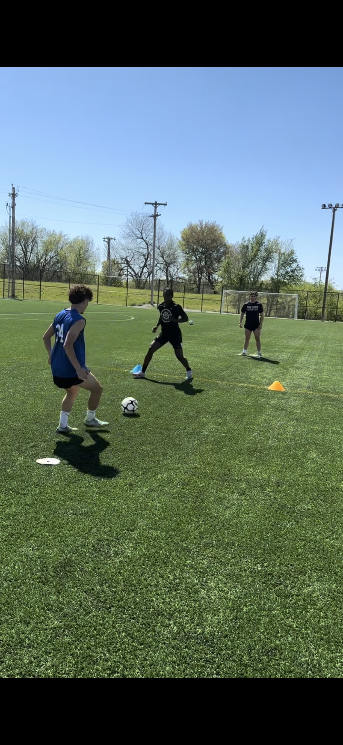 Soccer players training on a green field under a clear sky. One player in blue with ball, two others nearby.