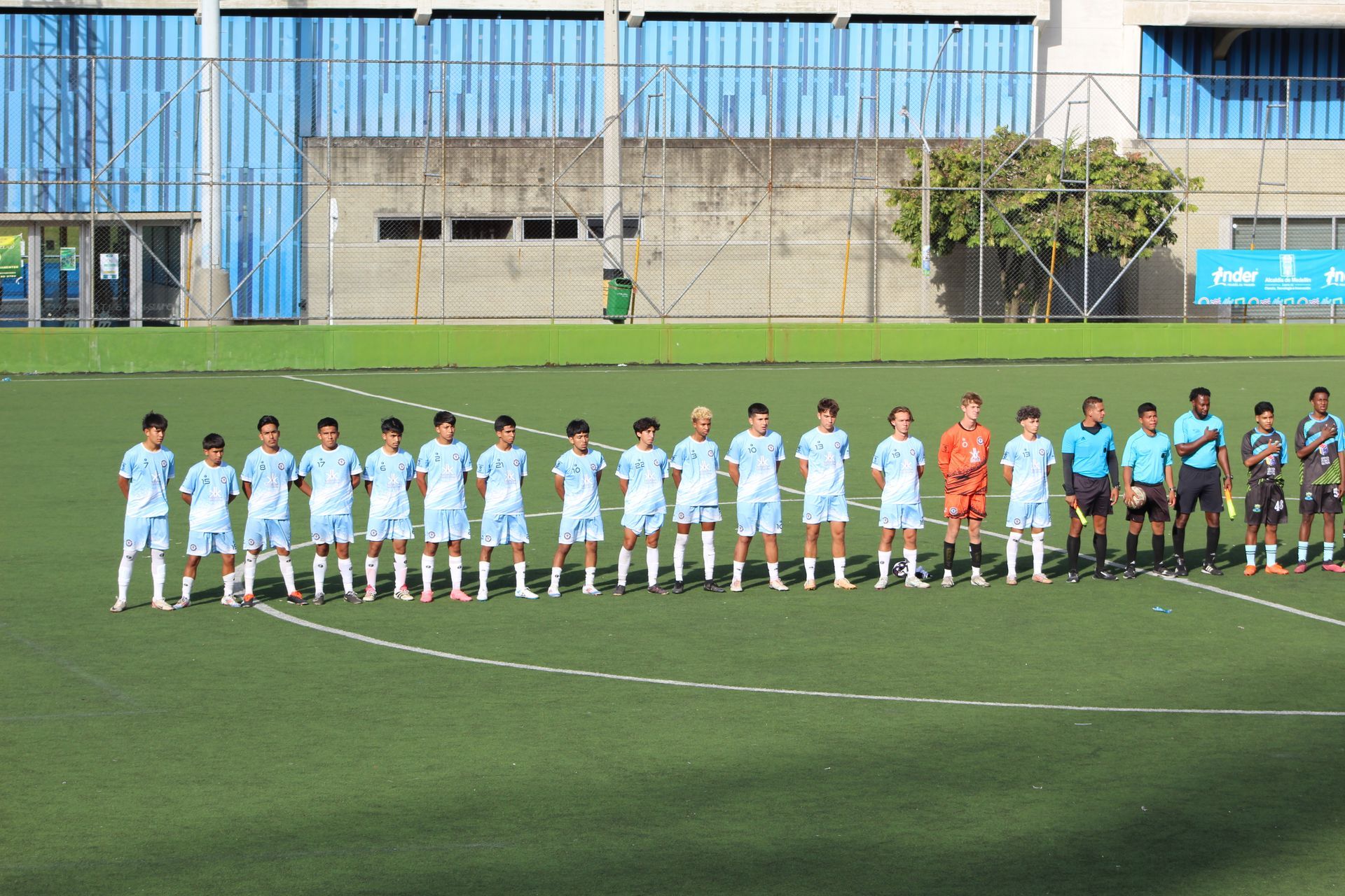 Soccer team in light blue jerseys on a green field, lined up for a team photo.