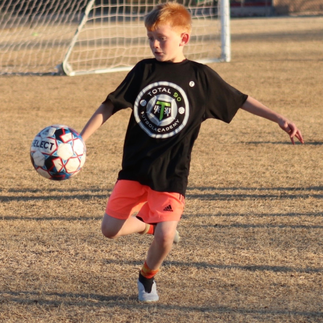Boy in black shirt and orange shorts, kicking a soccer ball on a field.