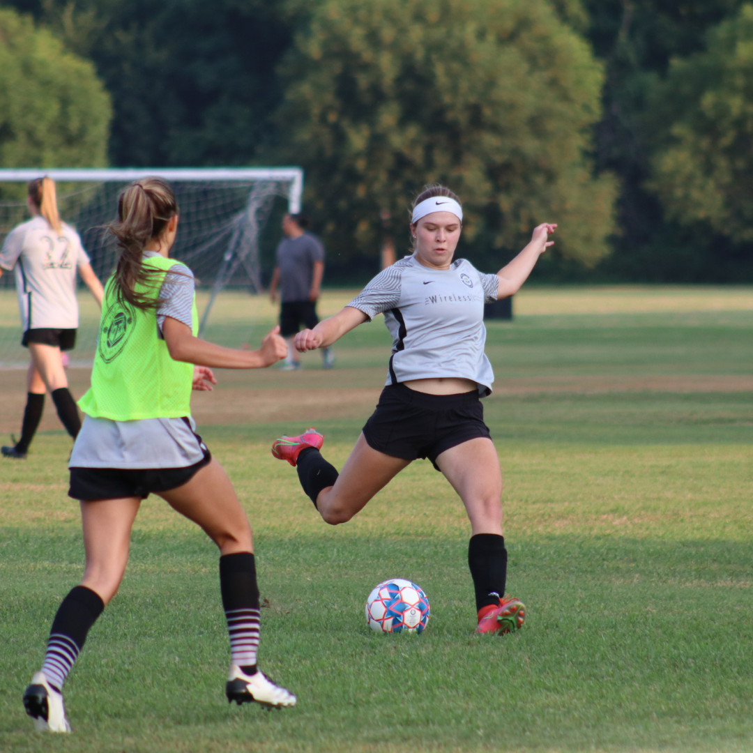 Woman in black shorts kicking a soccer ball on a green field, another woman in a neon vest watches.