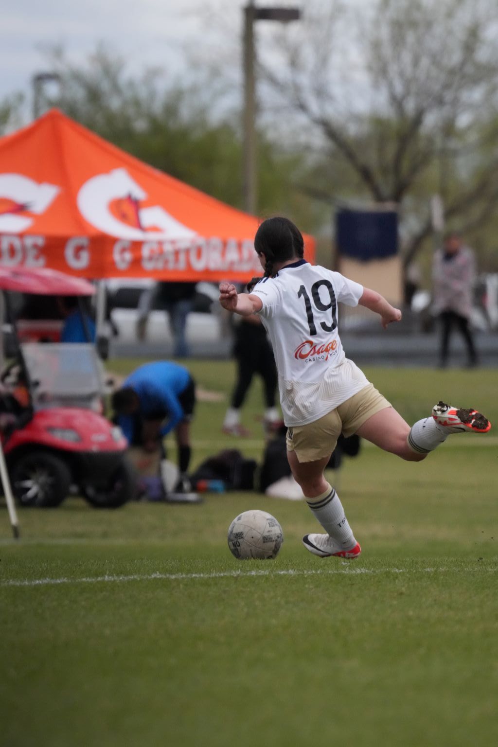 Soccer player kicking the ball on a green field; orange Gatorade tent in the background.