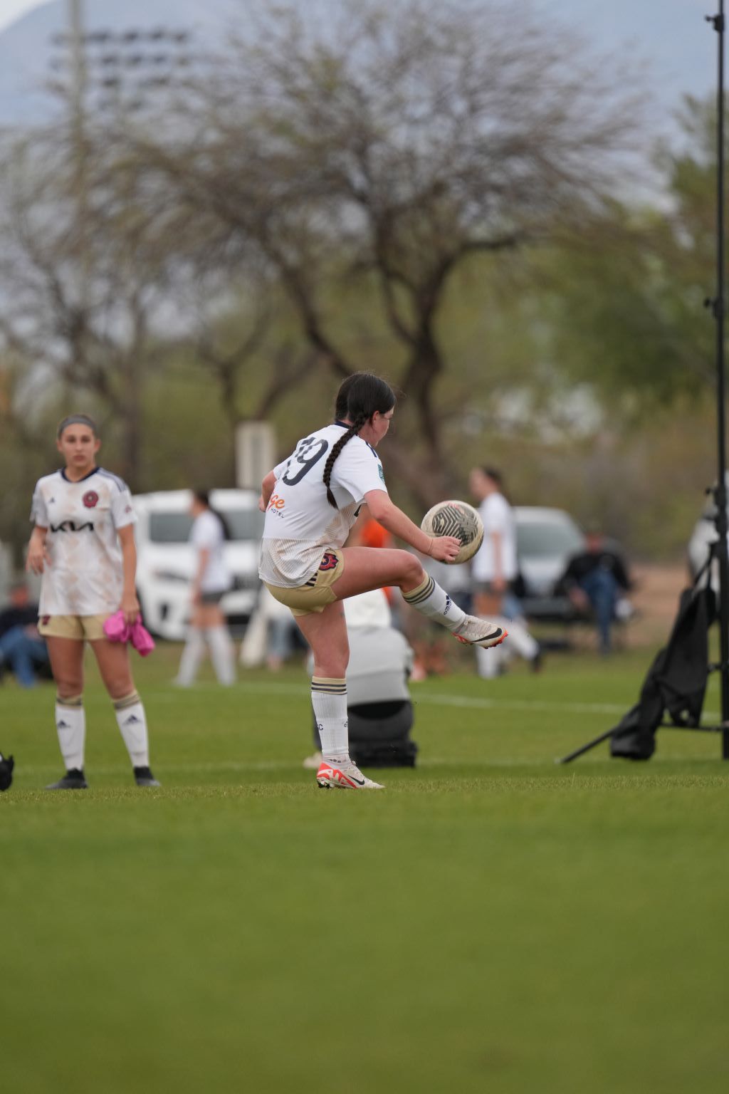 Woman in soccer uniform kicking ball on green field. Other players and parked cars visible.