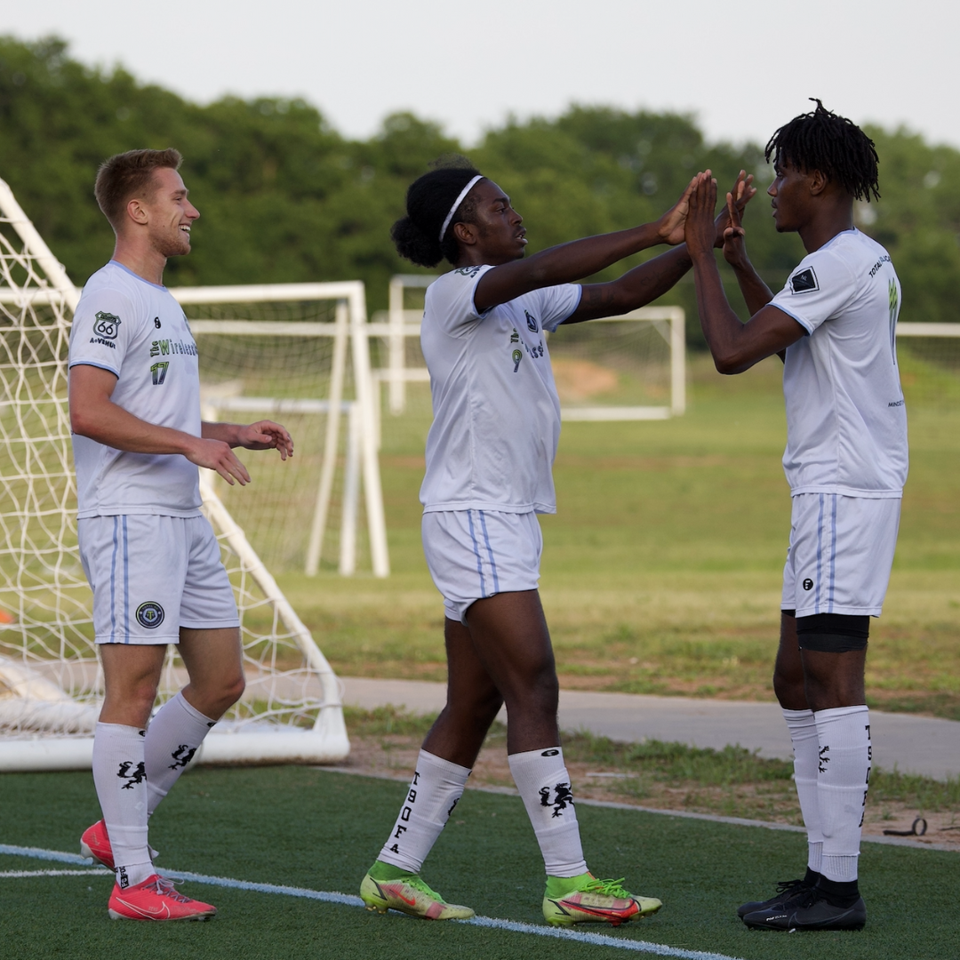 Soccer players in white uniforms celebrate on a field, giving high fives, near a goal.