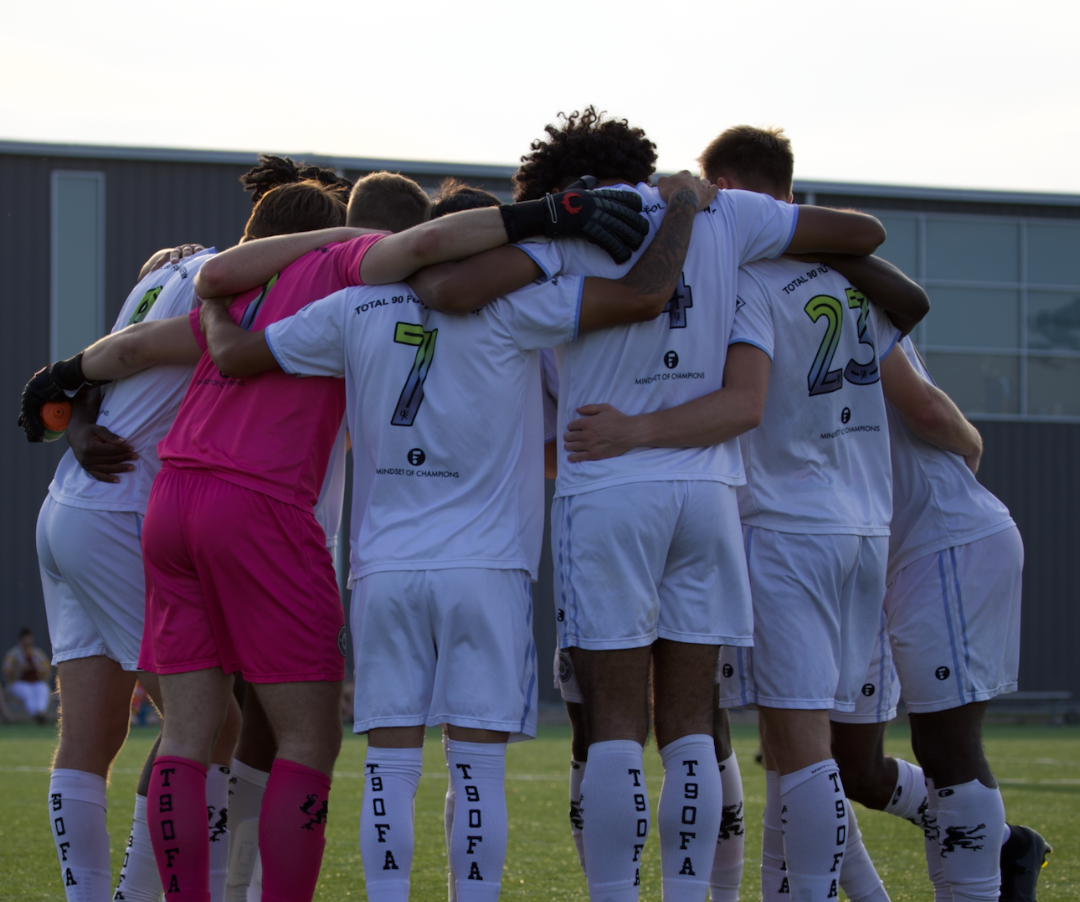 Soccer team huddling together with arms around each other, wearing white jerseys, pink goalkeeper.