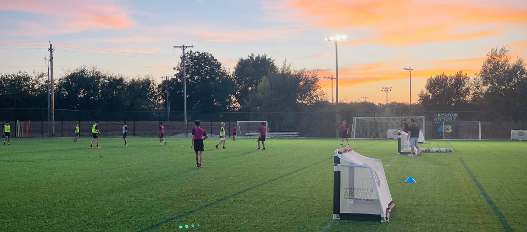 Soccer players training on a green field under a twilight sky, lights on.