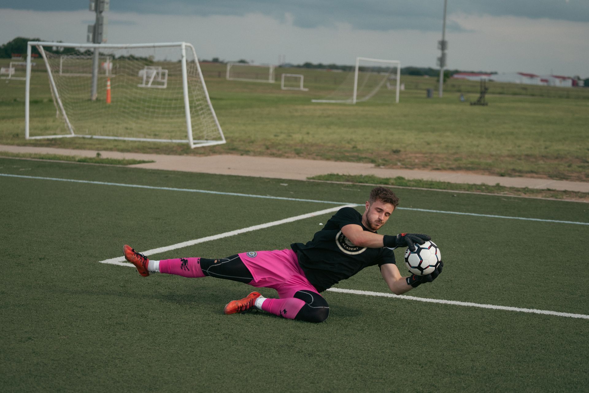Soccer goalie diving to catch the ball on a green field. He wears black shirt, pink shorts and socks.