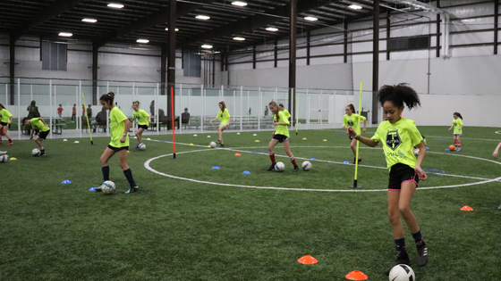 Girls in neon green soccer uniforms practice drills on indoor turf field.