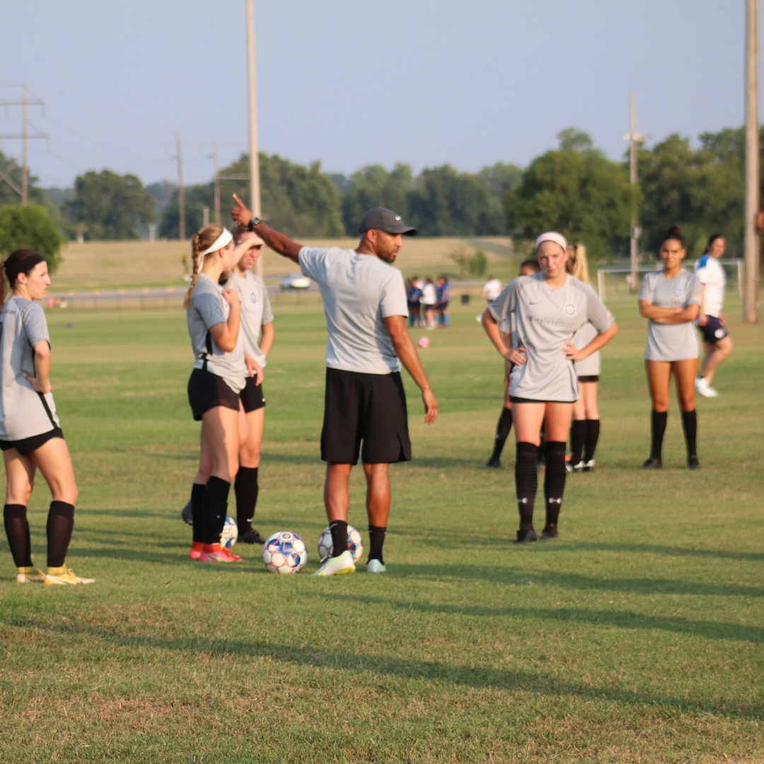Soccer coach with players on a field. Coach gestures, players listen. Sunny day.