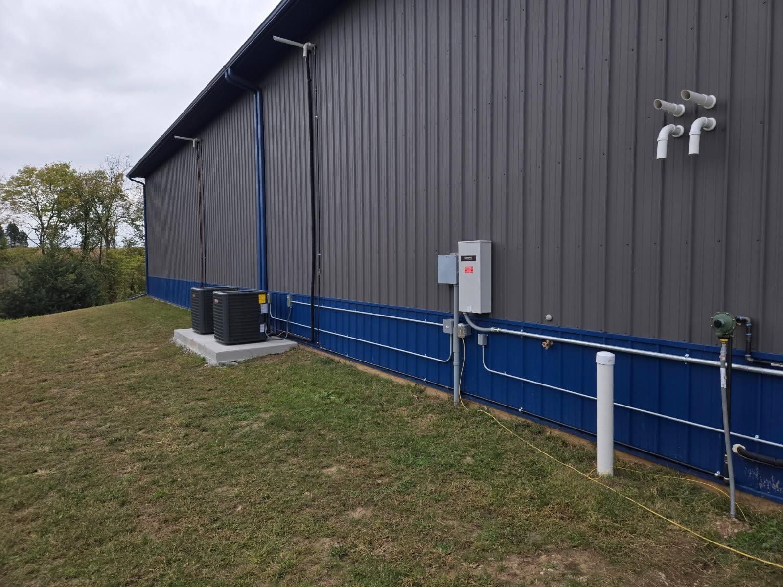 Side of a gray industrial building with blue trim, utility pipes, and electrical equipment beside a grassy lawn