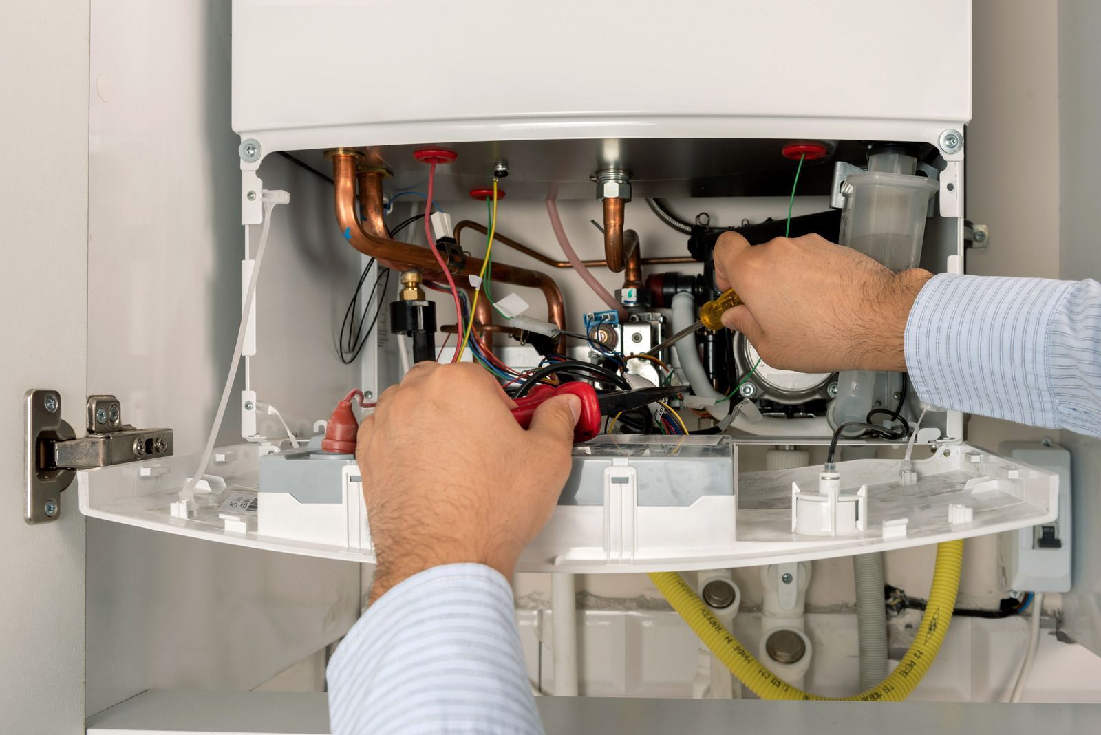 Hands repairing the interior of a wall-mounted boiler with tools and exposed wiring.