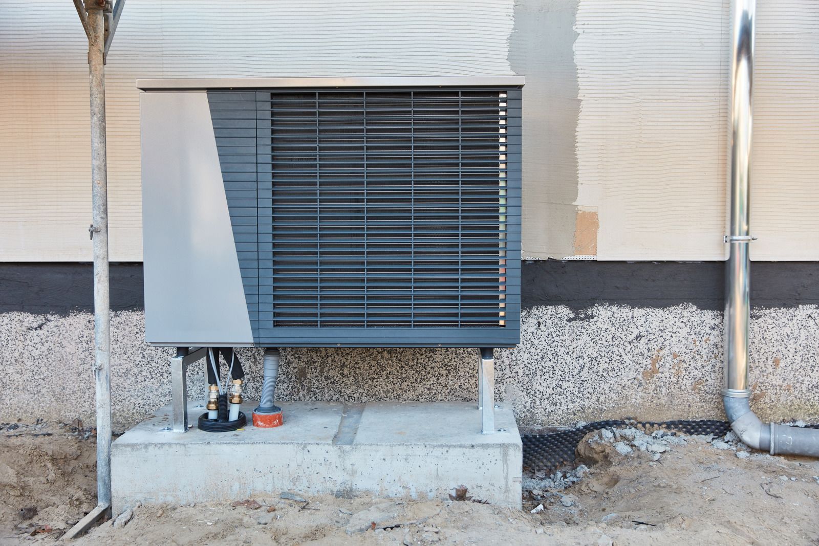 Outdoor wall-mounted HVAC unit with a gray vented cover and exposed pipes on a building exterior