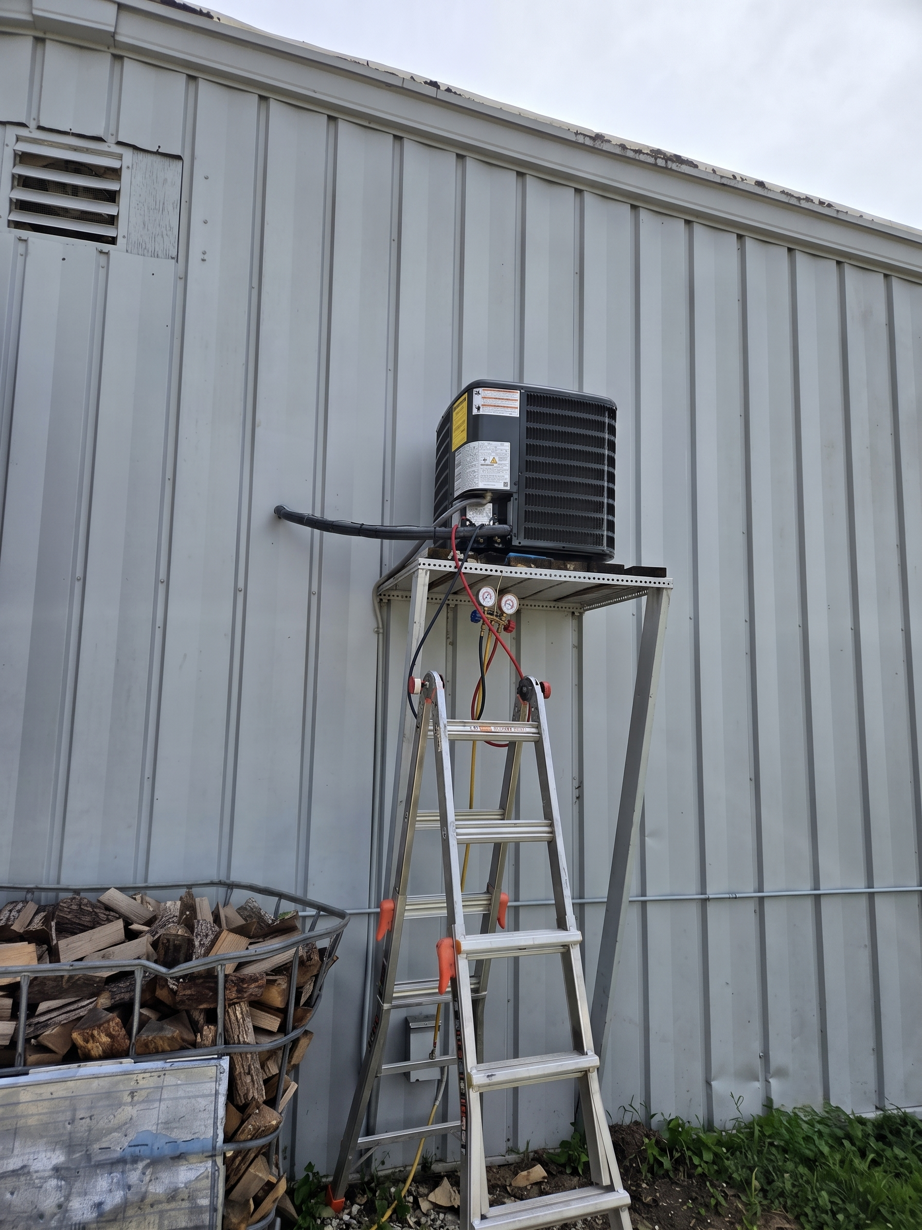 Ladder under a wall-mounted HVAC unit outside a metal building with firewood stacked nearby
