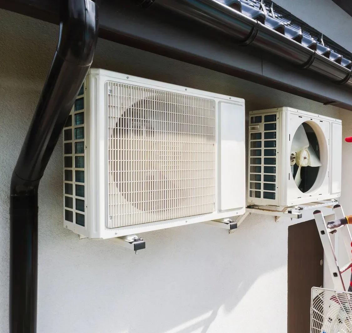 Two white outdoor air conditioner units mounted on a wall under a roof overhang.