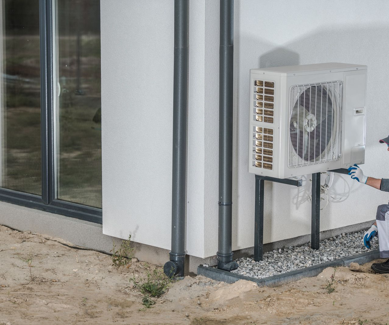 Exterior wall-mounted air conditioner unit beside a building with windows and exposed ground.